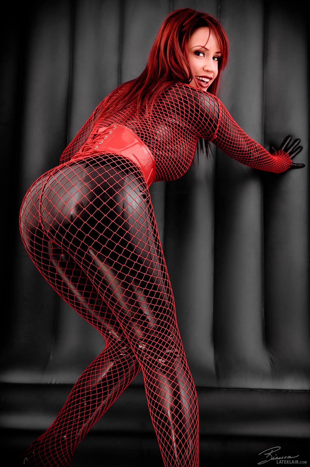ass bianca_beauchamp breasts catsuit corset female fishnets gloves large_breasts latex lipstick long_hair looking_back red_hair simple_background smile solo watermark