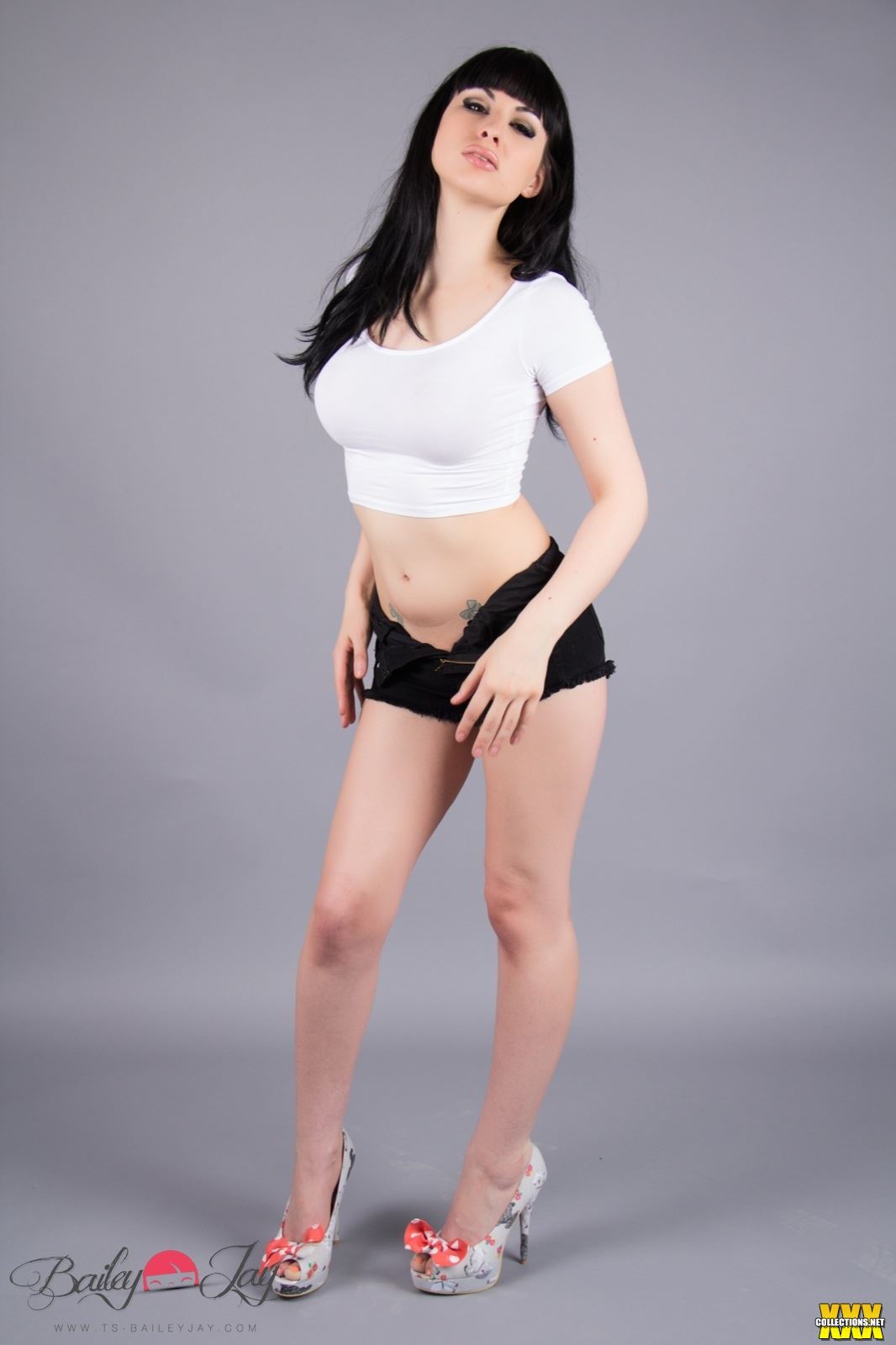 bailey_jay black_hair breasts clothed denim_shorts high_heels large_breasts long_hair looking_at_viewer midriff no_panties non-nude parted_lips porn_star shemale short_shorts shorts_undone simple_background solo tattoo transgender watermark white
