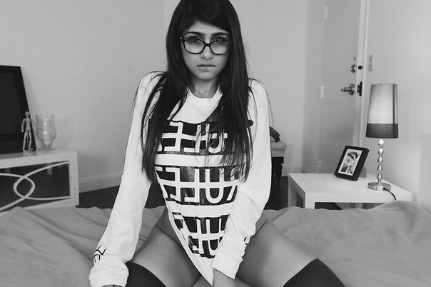 1girl biting_lip bottomless breasts brown_eyes brown_hair covering_pussy female female_only glasses greyscale improveme lebanese_american long_hair mia_khalifa monochrome non-nude on_bed partially_clothed porn_star shirt_pull sitting solo spread_legs stockings tan_skin thighhighs