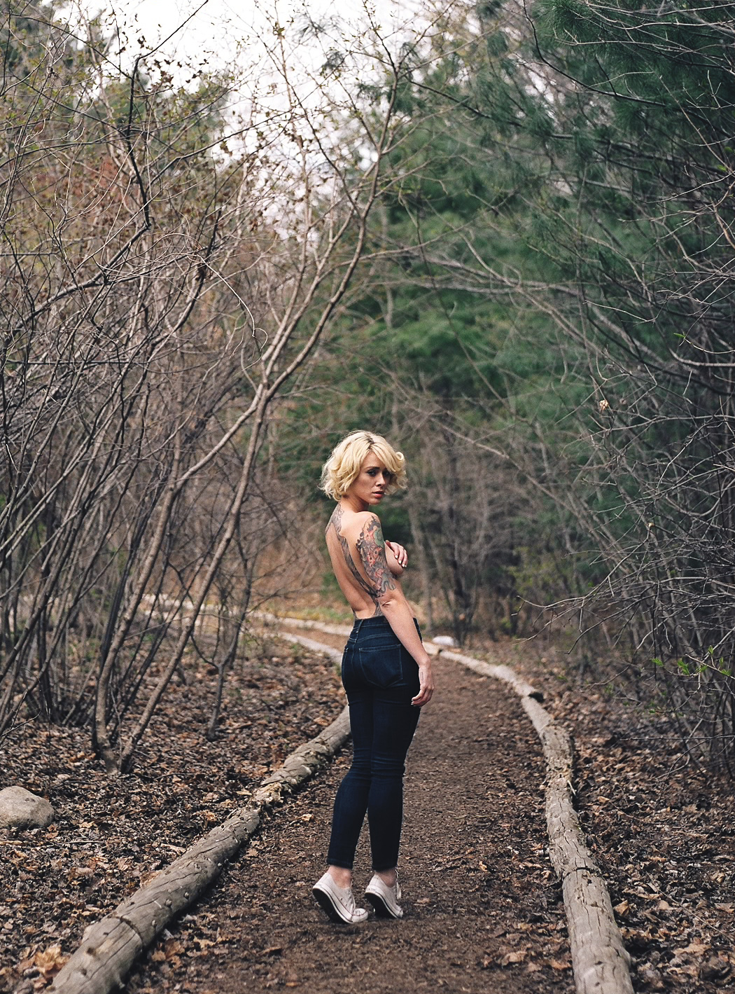 ass blonde_hair breasts covering_breasts denim female jeans looking_back sneakers solo tattoos topless woods