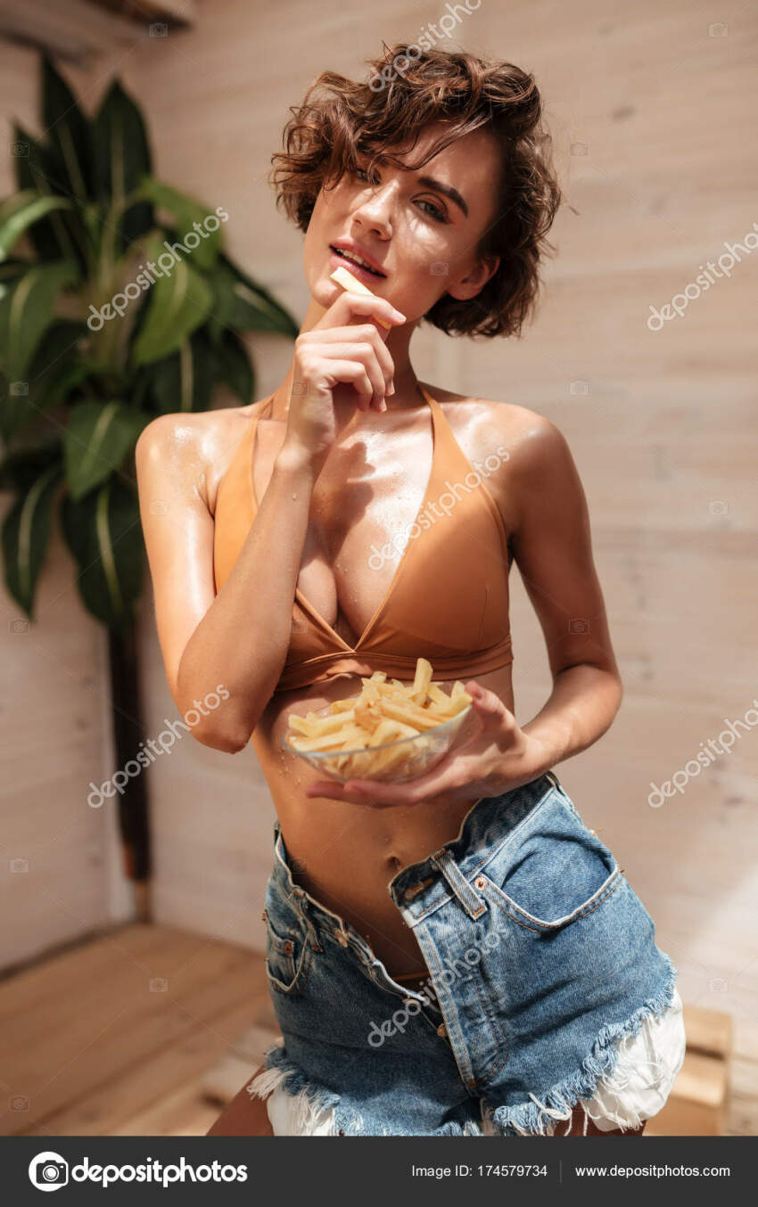 1girl bare_shoulders breasts brown_hair depositphotos eating female_focus food garetsworkshop jeans looking_at_viewer model navel one_eye_closed open_mouth orange_bra plant potatoes short_hair shorts sole_female solo standing stomach teeth watermark