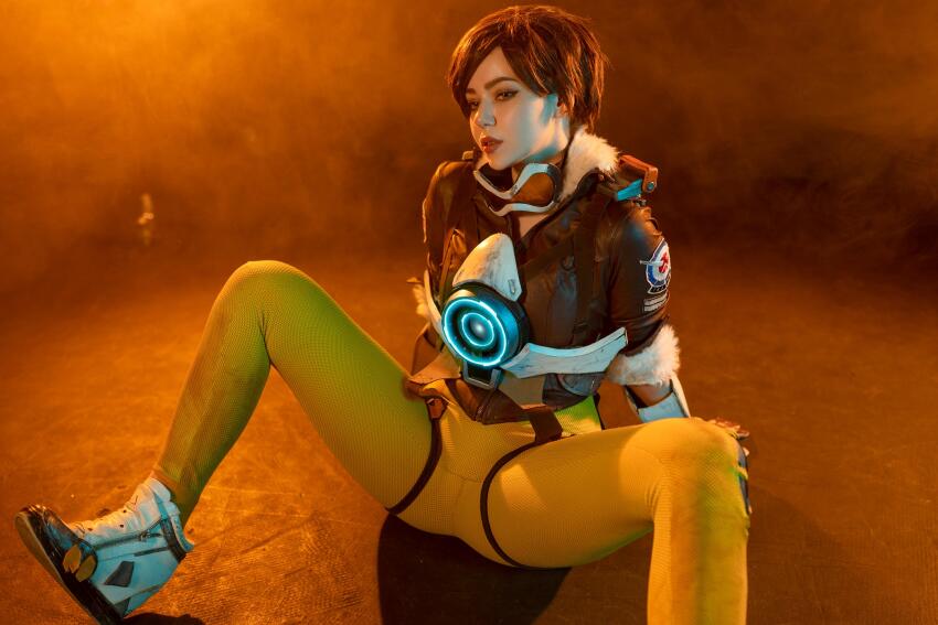 1girl alina_becker blizzard_entertainment brunette clothed clothed_female cosplay dark_hair dark_haired_female female_focus female_only form_fitting highres leggings non_nude on_floor overwatch photoshoot sexually_suggestive sfw skin_tight solo solo_female solo_focus spread_legs suggestive tight_pants tracer_(overwatch)